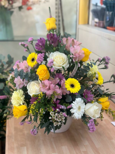 Pot of Joy - A lively and uplifting arrangement designed to brighten any space with its warm, cheerful palette. Featuring a beautiful blend of sunny yellows, soft pinks, creamy whites, and pops of purple, this design is artfully arranged in a modern container.