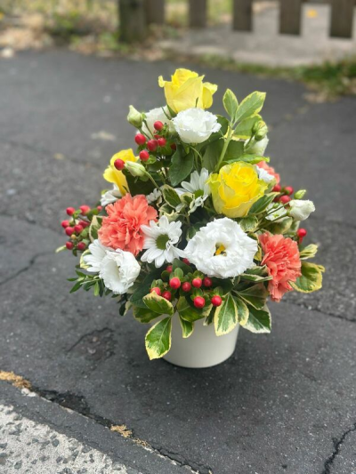 Sunny Charm Arrangement - A bright and cheerful arrangement featuring vibrant golden tones paired with soft coral hues, crisp whites, and pops of fresh greenery. Accented with glossy red details and variegated foliage, this design brings a lively, garden-fresh feel with plenty of texture and contrast. Arranged in a simple modern container, it’s perfect for uplifting any space, celebrating special moments, or sending a warm and joyful gift.