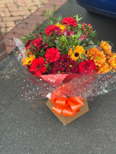 Fiesta Sunset - A bold and energetic bouquet bursting with rich reds, fiery oranges, and warm golden tones. Expertly wrapped in stylish presentation paper with a playful dotted finish and tied with a vibrant bow, this arrangement makes a striking statement. Lush greenery and textured accents add depth and movement, creating a lively, celebratory feel. Perfect for special occasions, congratulations, or sending a passionate and joyful gift.