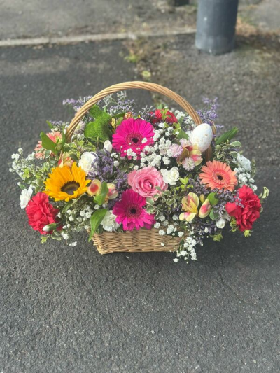 Countryside Celebration Basket - A joyful and vibrant basket arrangement inspired by a blooming countryside garden. Filled with bright pops of pink, sunny yellow, soft peach, and rich red tones, this design is full of life and cheerful energy. Delicate filler accents and lush greenery create a natural, textured look, while the woven basket and curved handle add a rustic, charming touch. Perfect for celebrations, gifting, or bringing a burst of color and happiness to any setting.