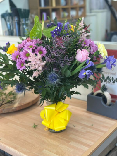 Radiant Garden Glow Vase - A bold and expressive arrangement bursting with rich purples, soft pinks, sunny yellow accents, and fresh greenery. Designed with a natural, free-flowing style, it features a mix of textures and striking shapes that create depth and visual interest. Presented in a clear vase and finished with a vibrant ribbon, this piece is perfect for celebrations, uplifting gifts, or adding a lively splash of color to any space.