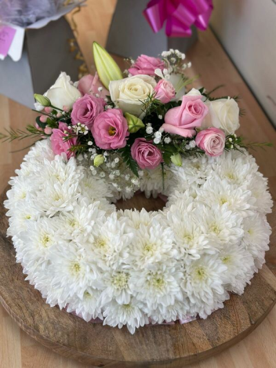 Massed wreath - A serene and elegant circular tribute arrangement designed to symbolize everlasting love and remembrance. Featuring a soft, pure white base accented with gentle blush and ivory tones at the top, this piece combines delicate textures with a refined, balanced design. The ring shape represents continuity and eternity, while the subtle greenery adds a natural touch. Ideal for memorials, sympathy gestures, or honoring a loved one with grace and beauty.