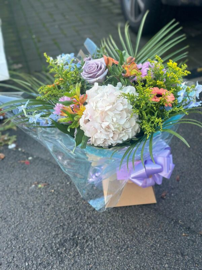 Refined Beauty - A fresh and uplifting bouquet featuring a soft blend of pastel tones, including gentle lilacs, creamy whites, warm peach accents, and touches of sunny yellow. Designed in a loose, airy style, it showcases flowing greenery and varied textures that create natural movement and depth. Finished with elegant wrap and a coordinating ribbon, this arrangement is perfect for celebrations, thoughtful gifts, or bringing a light and cheerful touch to any space.