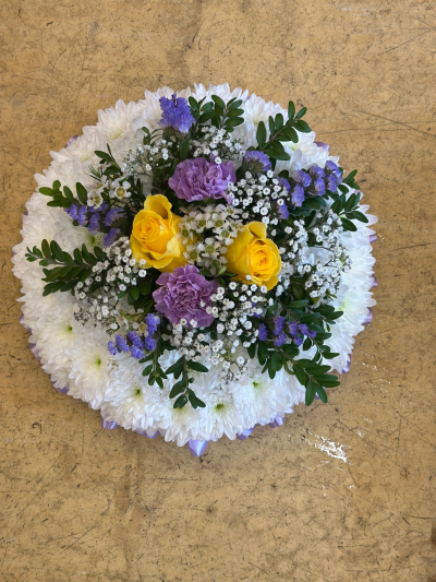 Massed Posy - A beautifully balanced circular tribute designed to symbolize unity, remembrance, and enduring love. Featuring a soft white base accented with touches of lavender, fresh greenery, and gentle pops of sunny color at the center, this arrangement offers both elegance and warmth. The layered textures and harmonious design create a peaceful and comforting presence, making it a thoughtful choice for memorials, sympathy gestures, or honoring a cherished life.