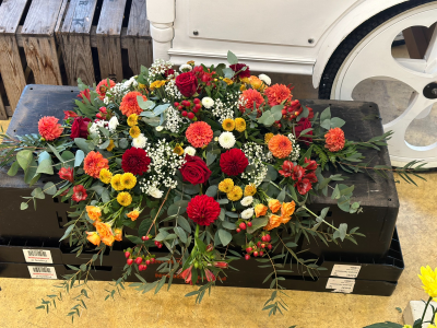 Autumnal Oasis Spray - A rich and vibrant tribute arrangement designed in a flowing, elongated style. Featuring a warm palette of deep reds, burnt oranges, golden tones, and fresh greenery, this piece captures a sense of warmth, remembrance, and celebration of life. Textured accents and trailing foliage create natural movement and depth, giving it an organic, garden-inspired feel. Perfect for memorial services or honoring a loved one with a bold and heartfelt expression.