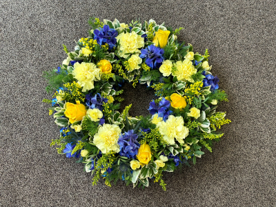 Sunlit Serenity Wreath - A beautifully balanced circular tribute designed to symbolise eternal love and remembrance. This vibrant wreath features a harmonious blend of golden and deep blue tones, creating a striking contrast that feels both uplifting and calming. Accented with fresh greenery and delicate textures, it offers a sense of peace and quiet strength. Perfect for honouring a life with warmth and dignity, this arrangement brings a gentle brightness to any memorial setting.