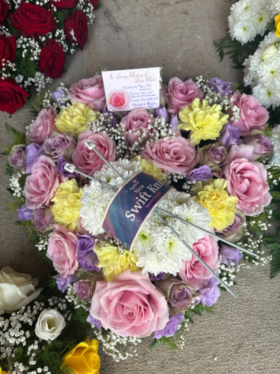 Knitting Tribute - A soft and heartfelt arrangement designed in delicate pastel tones, blending blush pinks, creamy yellows, lilac hues, and fresh whites. Thoughtfully arranged with fine textures and lush greenery, this tribute conveys tenderness, love, and cherished memories. The circular style symbolises eternity and continuity, while the gentle colour palette offers comfort and warmth. A beautifully meaningful piece, perfect for honouring a loved one with grace and affection.