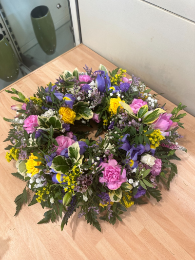 Pastel wreath - A beautifully arranged circular funeral wreath symbolising eternity, unity, and everlasting love. Designed in a soft yet uplifting palette of pastel tones, this tribute blends delicate textures with natural greenery to create a peaceful and comforting presence. The flowing, garden-inspired style reflects warmth, remembrance, and the celebration of a life well lived. A heartfelt and graceful tribute to honour someone dearly missed.